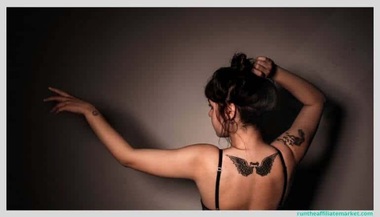 woman with dark hair and light skin posing with her back to the camera and pointing to her left. She has an intricate wing tattoo on her upper back