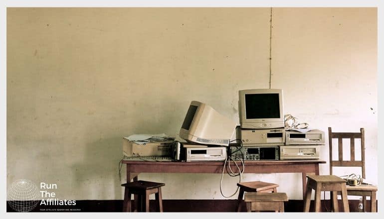 Old computer hardware and CRT monitors on a wooden desk with stools and chairs in the foreground